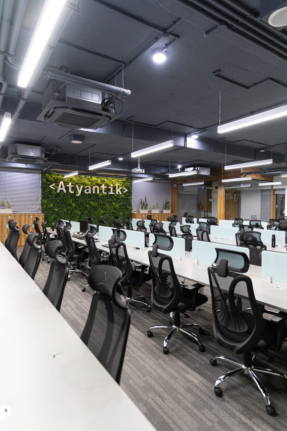Rows of empty desks with mesh chairs under linear ceiling lights, facing a living green wall carrying the Atyantik wordmark