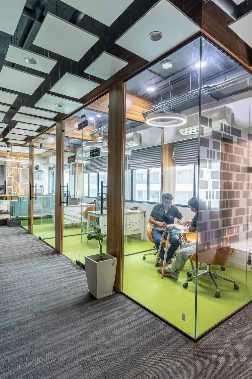 Two colleagues review printed work at a round table inside a glass-walled meeting pod