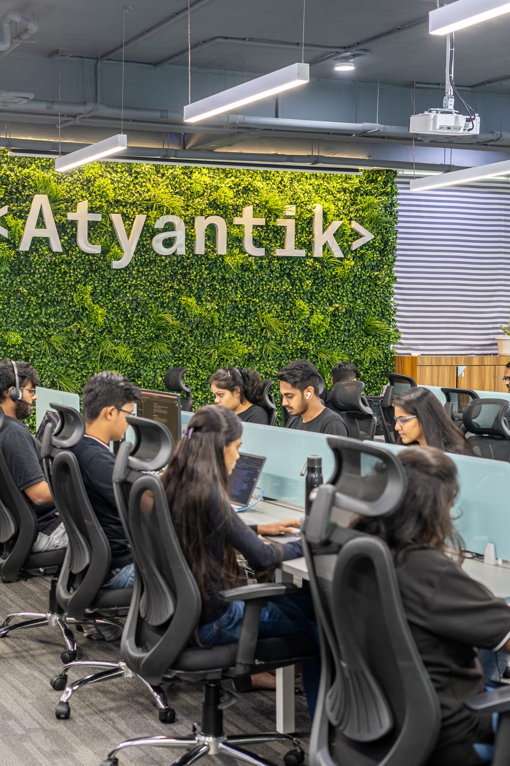 Software engineers working at laptops along a shared desk in front of the moss wall spelling Atyantik